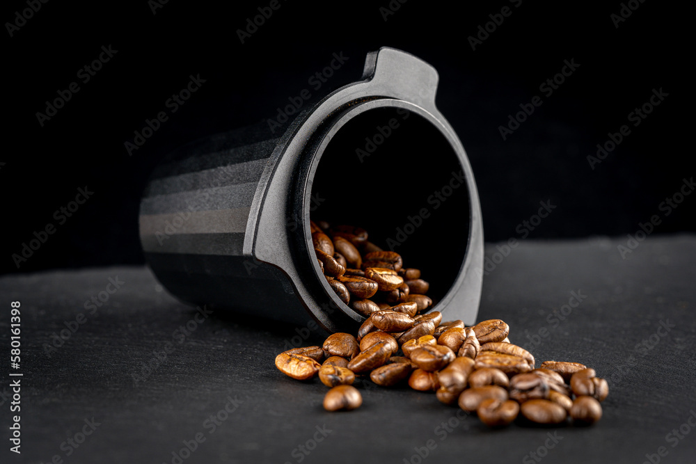 Dosing cup with black matte texture. Top view on black background with coffee beans. Barista tool for dosing coffee beans and using it to when grinding beans. Focus on the beans in the center..