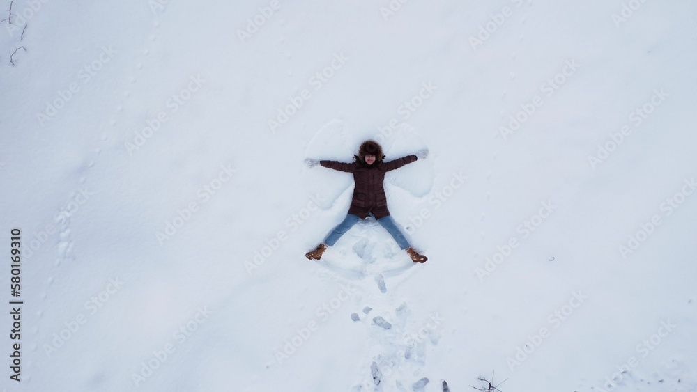 A young woman lies on the snow and makes the figure of an angel. View of the girl from above. The camera moves away.