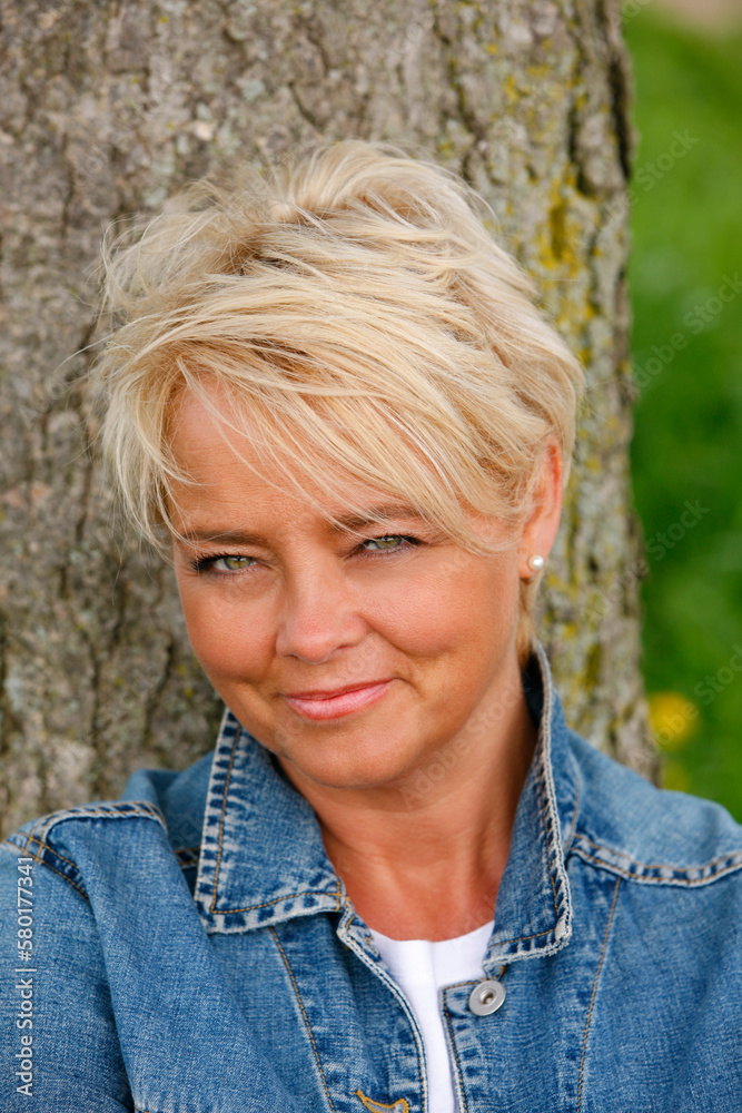 Close up portrait of attractive mature blond woman leaning against tree outdoors.