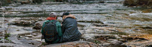 Rear of couple of hikers sitting on bank of rocky river and resting when hiking in mountains.