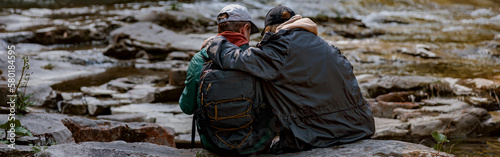 Back view of couple of hikers sitting on bank of rocky river and hugging. Hiking in mountains.