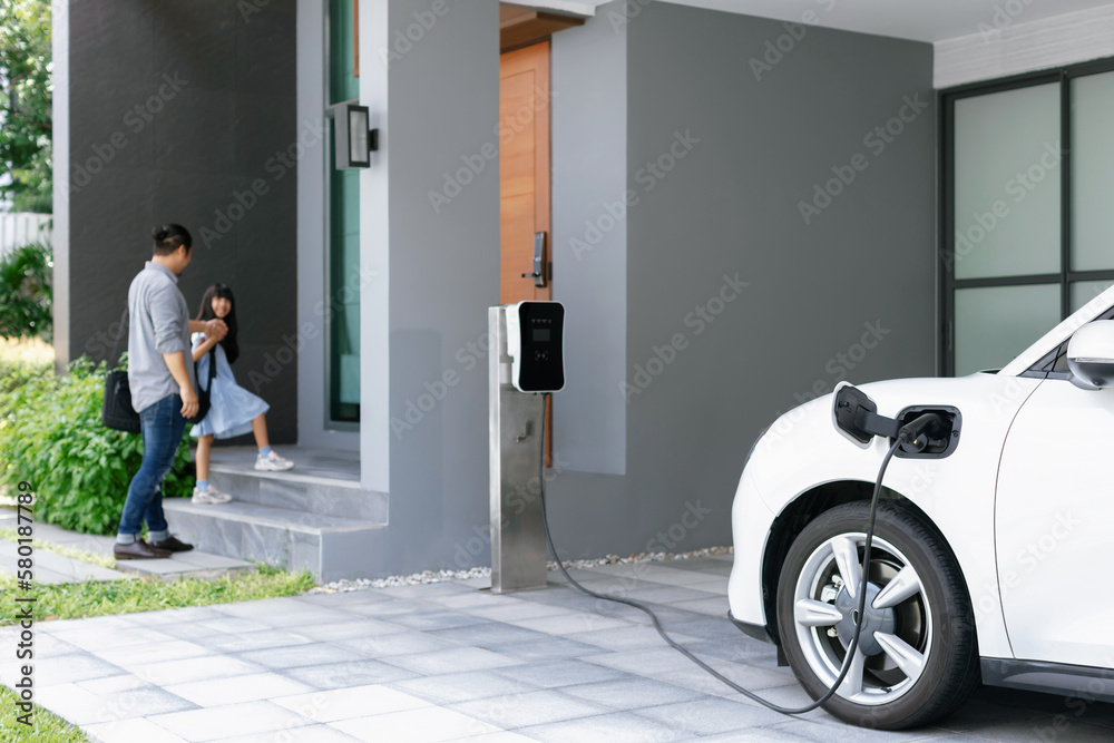 Focus progressive electric vehicle recharging at home charging station using clean and renewable energy with blurred father and daughter walking in background for future renewable energy concept. - obrazy, fototapety, plakaty 