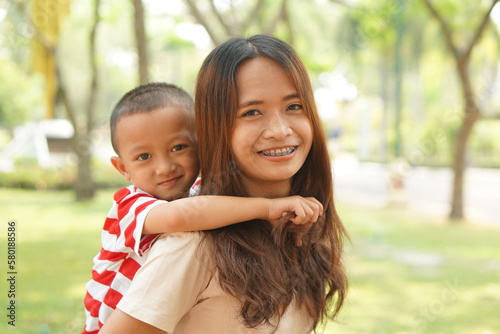 A boy happily rides on his mother's back in the park.