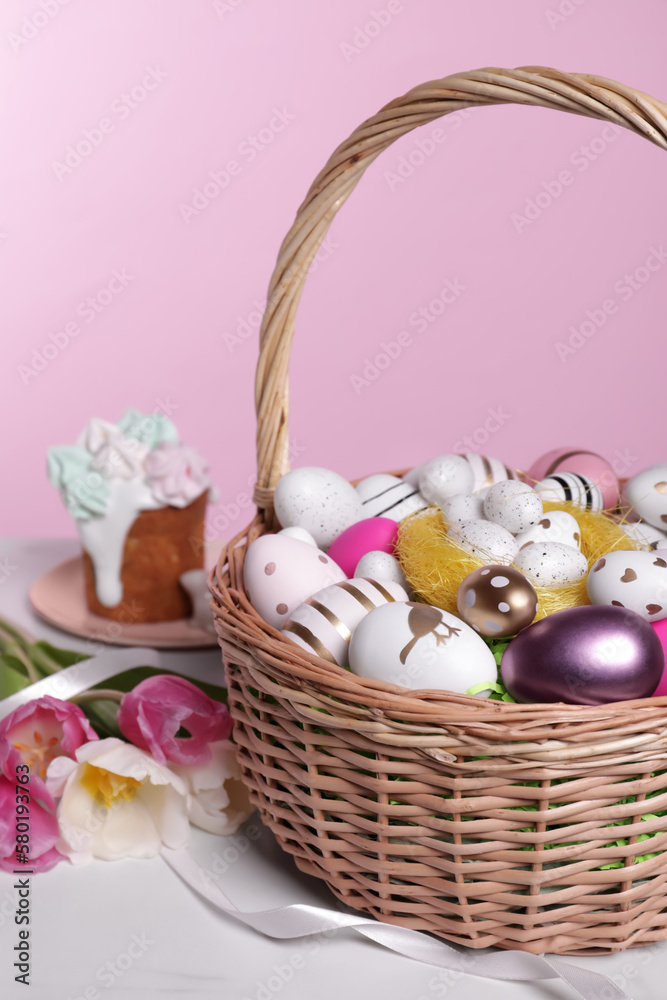Wicker basket with festively decorated Easter eggs and beautiful tulips on white marble table against pink background
