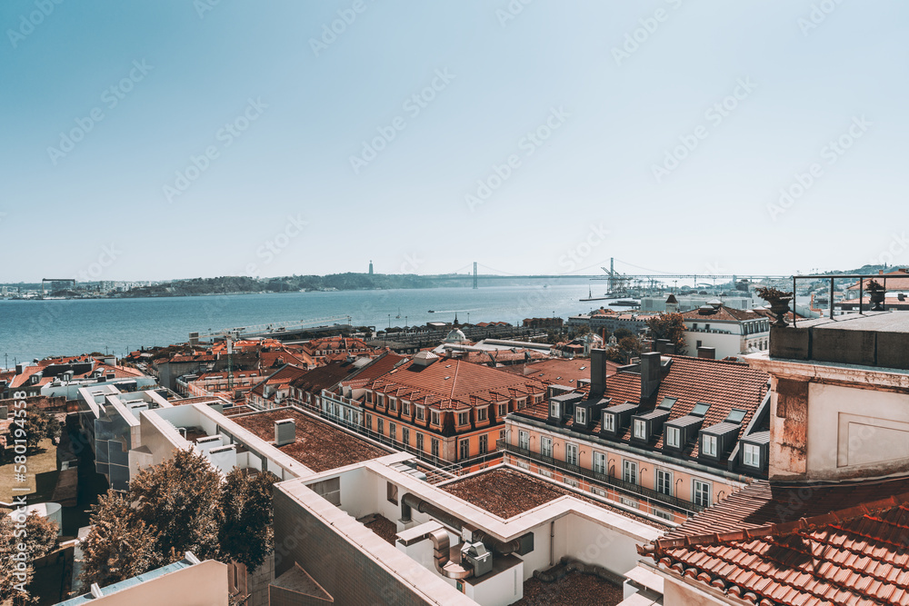 A sunny shot of Lisbon's city center, a cluster modern design buildings, with red brick roofing, for housing, and local businesses, and in the background the 25th of April Bridge can be seen far away