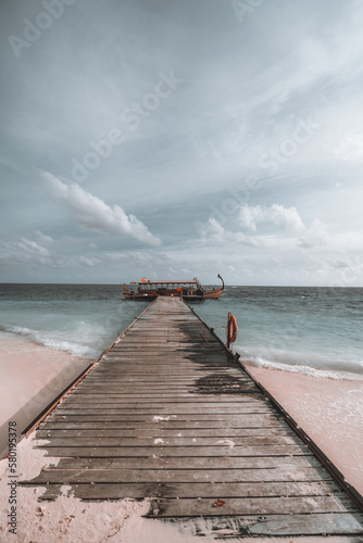 A vertical shot of a very long wooden pier on a Maldivian beach island, leading to a simple yet beautiful gondola boat moored in the grey seawater on a day with clouds in the sky