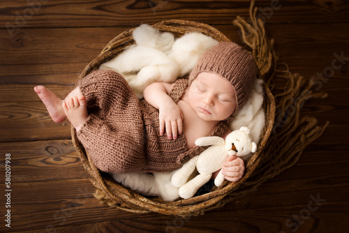 Top view of a newborn baby sleeping in a brown overalls, in a brown knitted cap, on a white felt flokati. On a dark wooden background. Beautiful portrait of a little newborn 7 days, one week old. photo