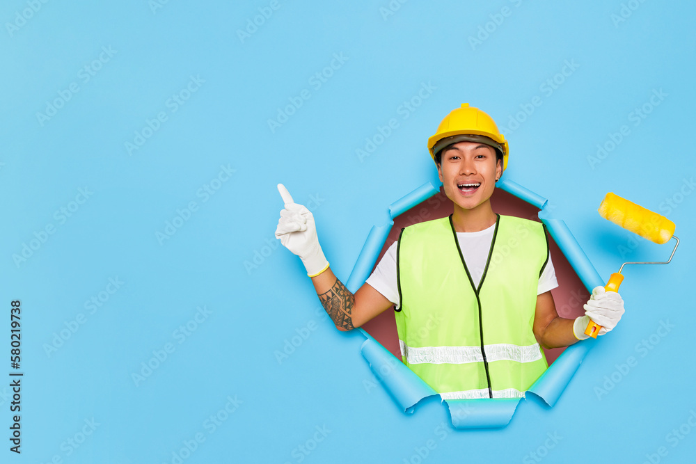 Indoor shot of positive Asian male builder with pain roller in a hand feels excited ready for house renovation wears hardhat protective gloves and uniform pointing index finger aside to the copy space Indoor shot of positive Asian male builder with pain roller in a hand feels excited ready for house renovation wears hardhat protective gloves and uniform pointing index finger aside to the copy space