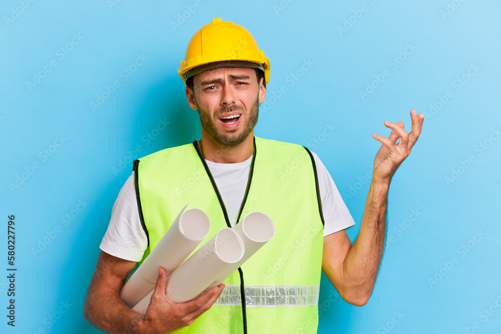 Young construction engineer holds blueprints with confused face expression isolated over blue background Young construction engineer holds blueprints with confused face expression isolated over blue background