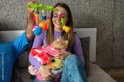 A young couple on the bed hugs their cat wrapped in a baby blanket and plays with her with a baby rattle. Conscious refusal of a couple to have children, treatment of pets as children. photo