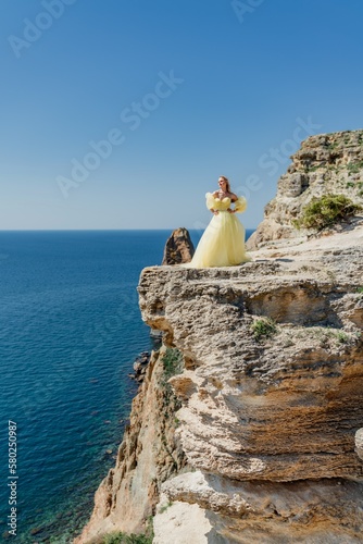 Woman in a yellow dress on the sea. Side view Young beautiful sensual woman in yellow long dress posing on a rock high above the sea at sunset. Girl in nature against the blue sky