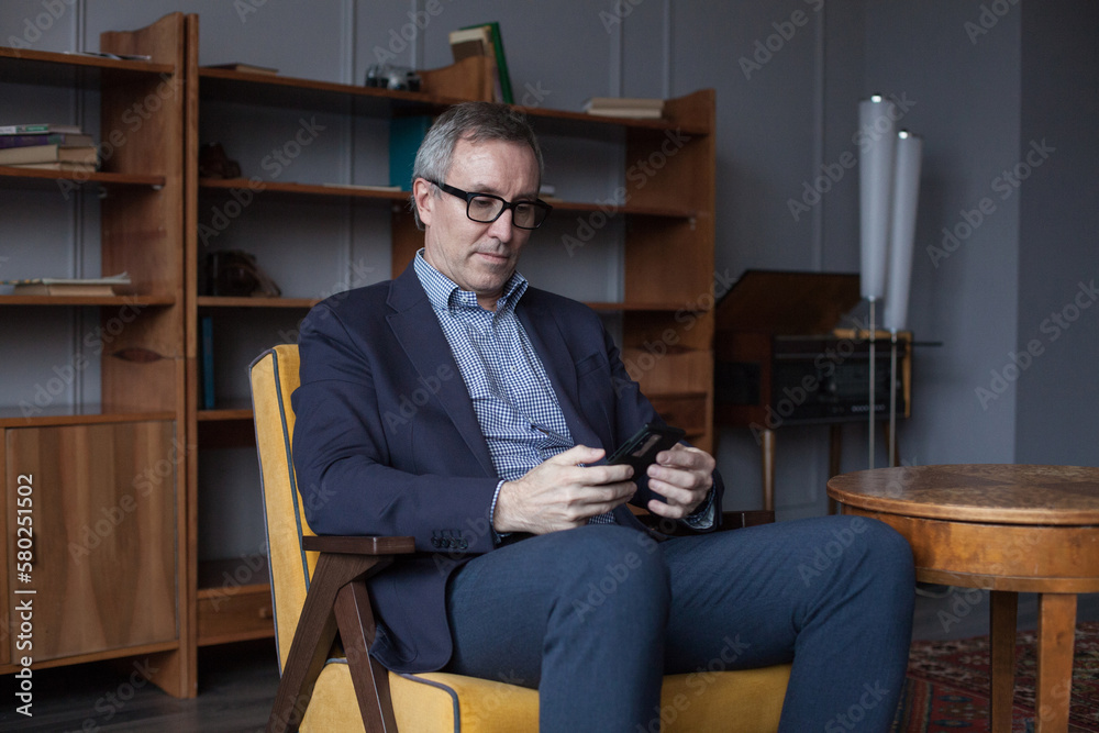 Confident elderly businessman with grey hair in suit and glasses working and typing on mobile phone indoors on background of office wall with boockshelf