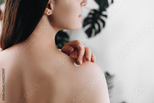 Close up back view of young female 20s old years applying cream on shoulder standing in cozy light bathroom at home. Beautiful lady apply body lotion. Health and beauty concept. Daily skincare routine