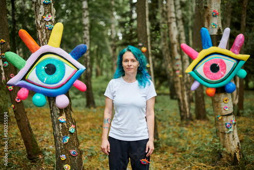 Weird young adult woman with dyed turquoise hair in white T shirt near big eyes art installation in outdoor art exhibition in public park. Female handmade artist near colorful eyes art objects