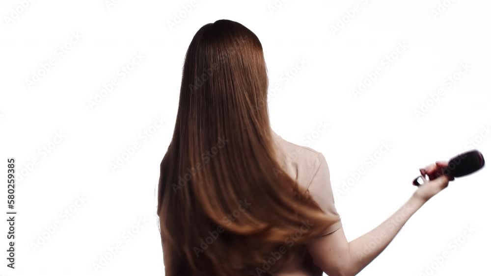 Woman in a pink t-shirt slowly combing her long healthy shiny brown hair with a comb in a back view on a white background. Woman getting her hair done. Hair care concept