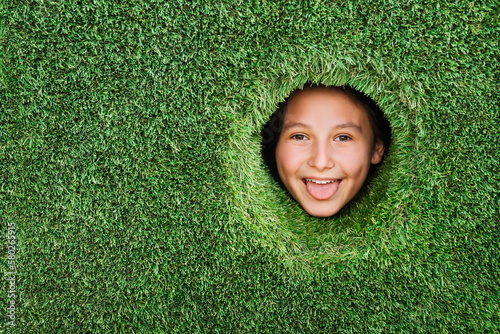 Advertise lawn and artificial green grass. Little kid with funny face looks into round hole in a wall made of green grass. Green space, eco-friendly, organic materials concept. photo