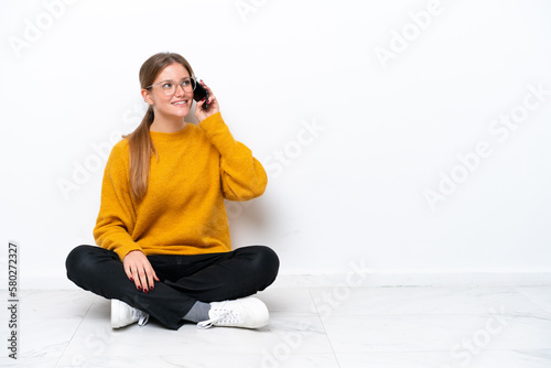 Young caucasian woman sitting on the floor isolated on white background keeping a conversation with the mobile phone