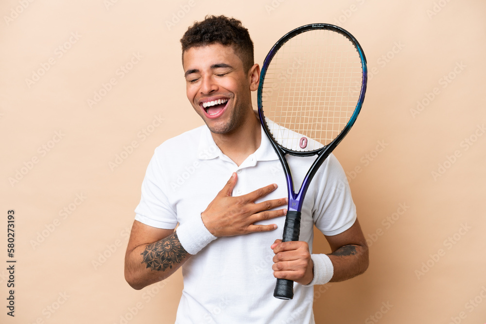 Young brazilian handsome man playing tennis isolated on beige background smiling a lot