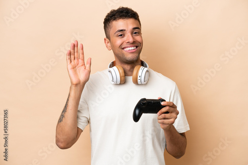 Young Brazilian man playing with a video game controller isolated on beige background saluting with hand with happy expression