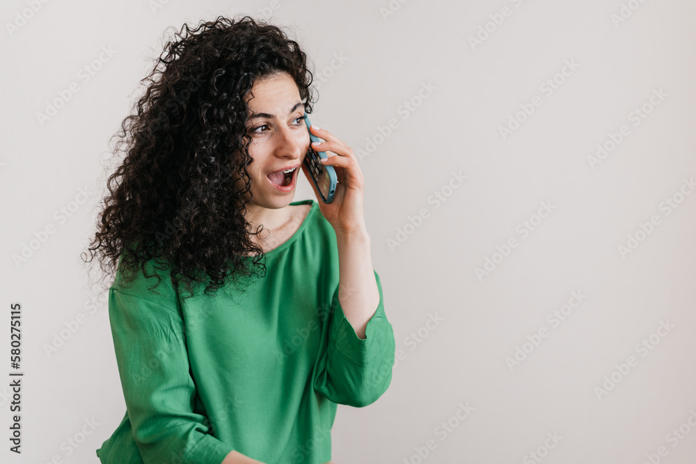 Excited Spanish young adult woman in green blouse talking by phone with opened mouth astonished by reecived news, gossip. Curly brunette business woman against grey wall with copy space. Excited Spanish young adult woman in green blouse talking by phone with opened mouth astonished by reecived news, gossip. Curly brunette business woman against grey wall with copy space.