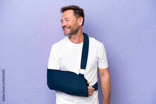 Middle age caucasian man with broken arm and wearing a sling isolated on purple background looking side