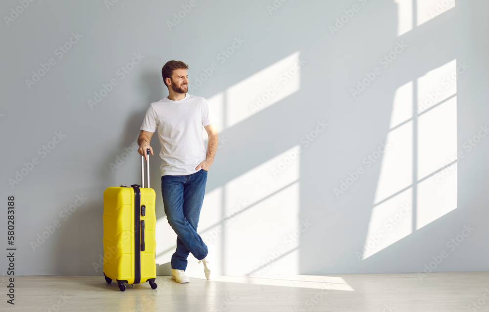 Full body length shot of young man with yellow suitcase, in white T shirt and jeans standing by grey wall background and looking aside at blank copy space. Travelling, holiday, vacation trip concept Full body length shot of young man with yellow suitcase, in white T shirt and jeans standing by grey wall background and looking aside at blank copy space. Travelling, holiday, vacation trip concept