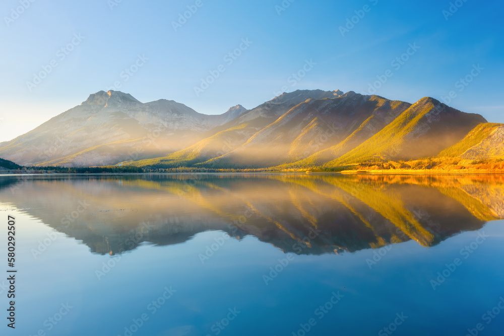 Lake and mountains in a valley at dawn. Reflections on the surface of the lake. Mountain landscape at sunrise. Foggy morning. Natural landscape with bright sunshine.