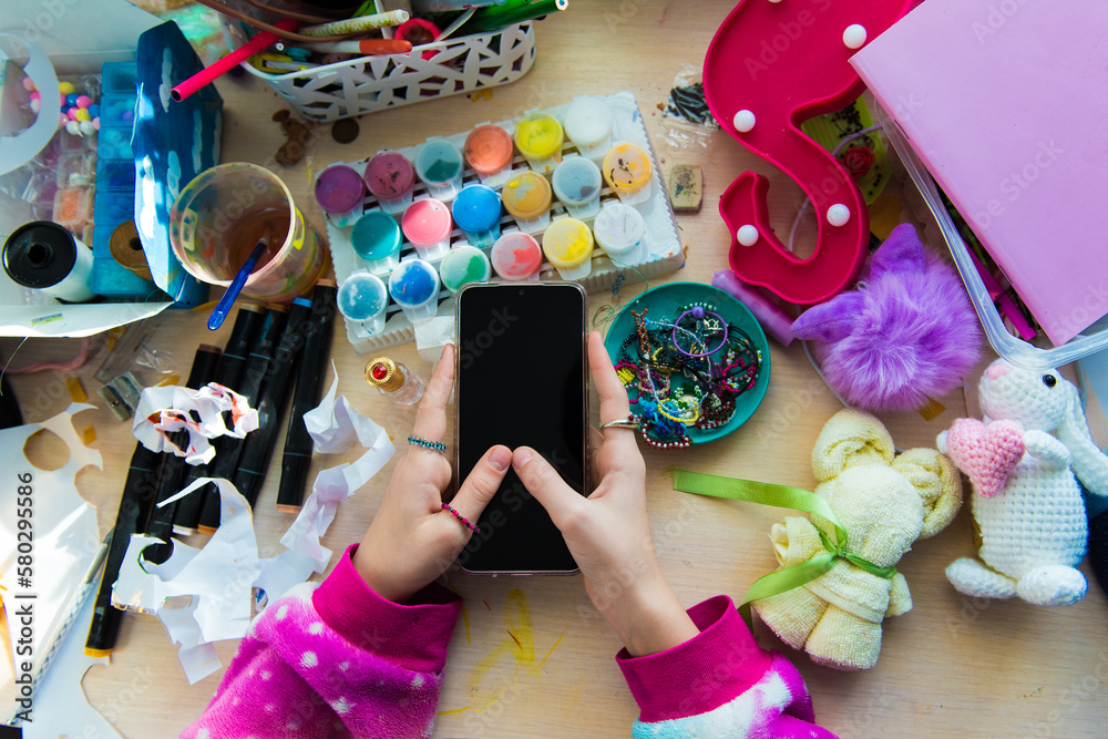 Obraz premium Hand of child girl using smartphone on chaotic desk in home office. Desk with objects for creativity, paints for drawing, children toys randomly scattered