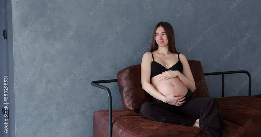 pregnant woman sitting on a brown sofa smiling and holding her big bare belly. pregnant woman sitting on a brown sofa smiling and holding her big bare belly.