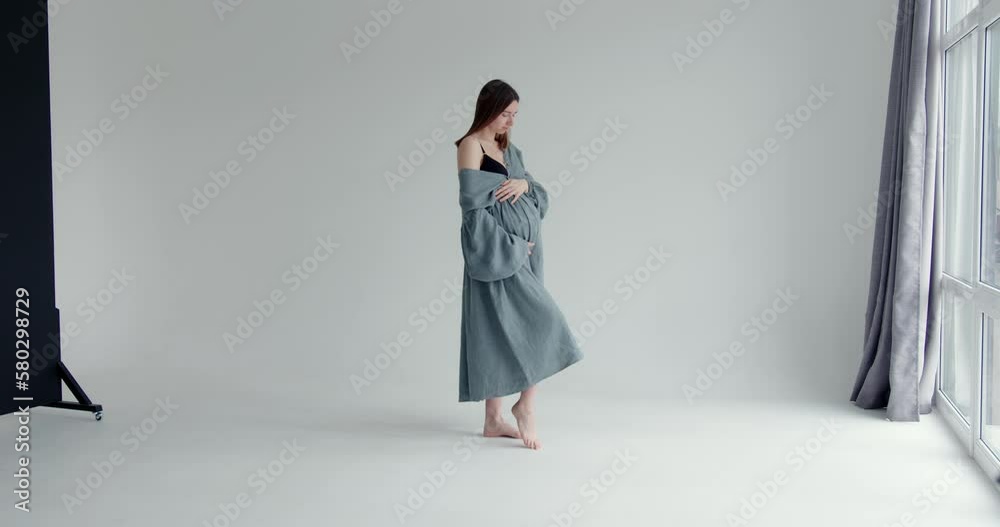 beautiful pregnant woman in a long dress stands in the studio on a white background near the window and holds her belly. general plan beautiful pregnant woman in a long dress stands in the studio on a white background near the window and holds her belly. general plan
