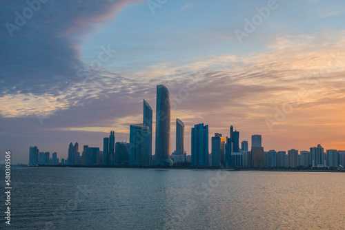 Abu Dhabi cityscape at sunrise with clouds and water in front.