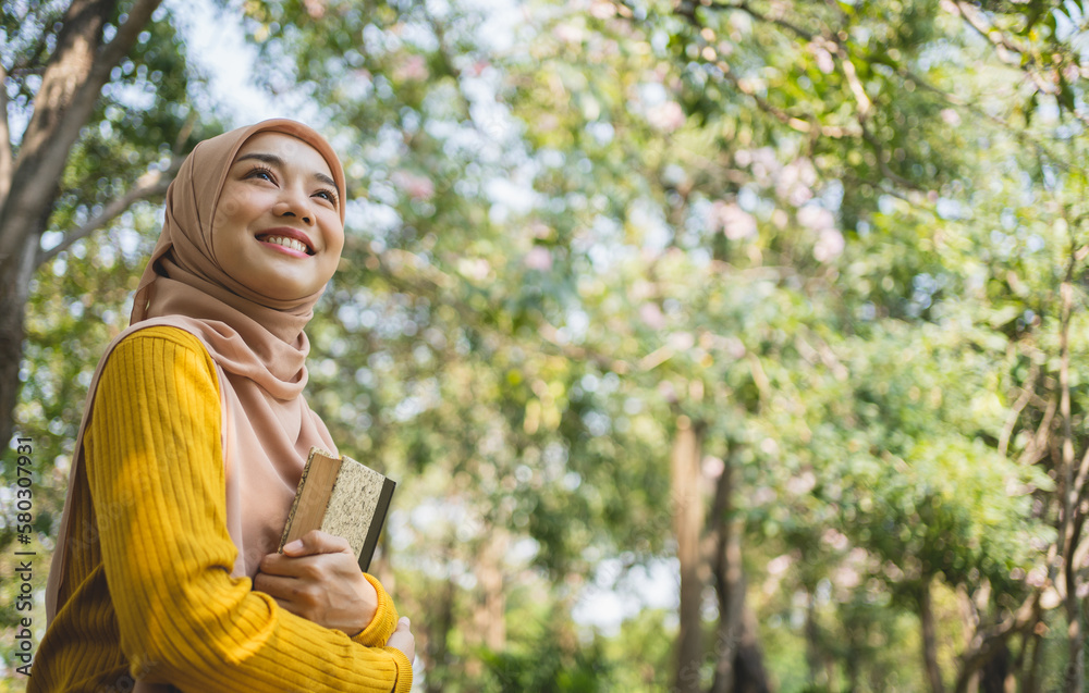 Portrait of a delighted young Asian Muslim girl sitting on a grass at the park, reading a book.