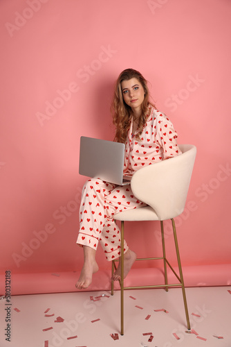 beautiful woman in heart print pajamas on a pink background is working on a laptop. Pink pajamas with red hearts. clothes for sleep and home. Valentine's Day. work at the computer in pajamas at home