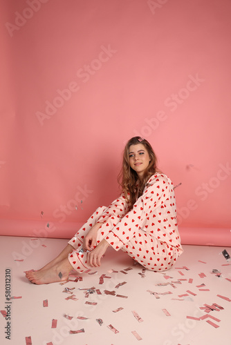 beautiful girl in heart print pajamas on a pink background. throw festive confetti. Pink pajamas with red hearts. clothes for sleep and home. Valentine's Day. beautiful blonde in pajamas
