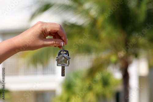 Fototapeta Naklejka Na Ścianę i Meble -  Real estate agent, home keys in female hand on background of house surrounded by palm trees. Buying or rental a villa on ocean coast, removal to tropical country