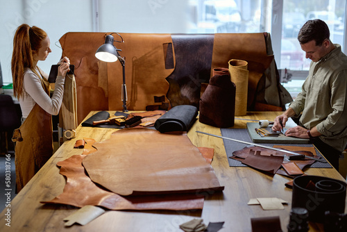 ginger woman in apron using stitching clamp at work, her colleague making holes on leather material with awl. side view shot