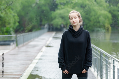 Portrait of young beautiful female blond hair crossed arms in black coach jacket standing in nature park looking at camera.