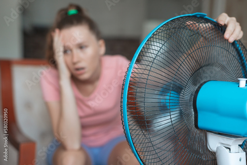 Young woman using electric fan at home in living room, sitting on couch cooling off during hot weather, suffering from heat, high temperature photo
