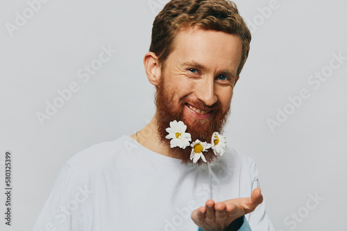 Photography Portrait of a funny man in a white T-shirt with flowers daisies in his beard on a white isolated background, copy place