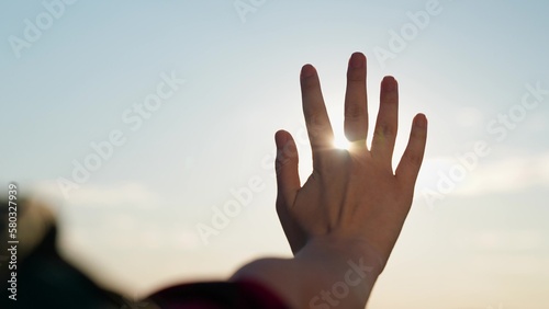 Happy teenage girl touches sun with her fingers. Hand of happy young woman plays with glare of sun. Girl holds out her hand at sunset. Sunrise between womans hands. Dreams of young woman traveler