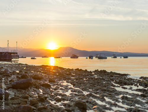 Morning atmosphere at Lake Garda. The water level is historically low, so rocks and stones on the shore stand out. In the background the water, boats, buoys and the shore in the morning light