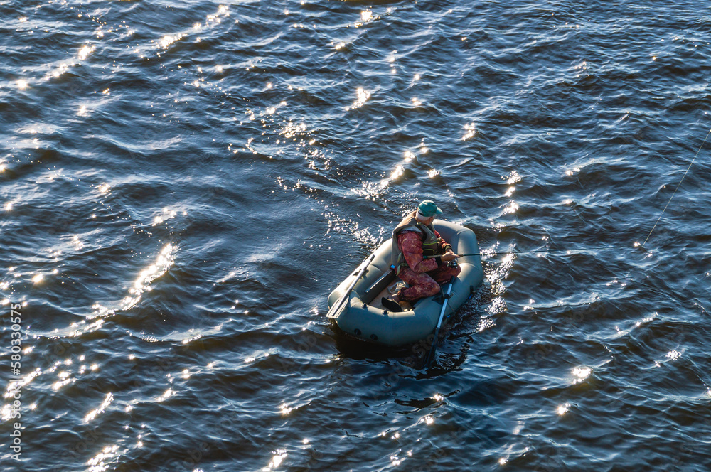 Fototapeta premium A man is fishing on a spinning in a rubber inflatable boat. Fishing on the river. Fishing with a fishing rod. Rest on the river. A fisherman catches fish while in a rubber boat.