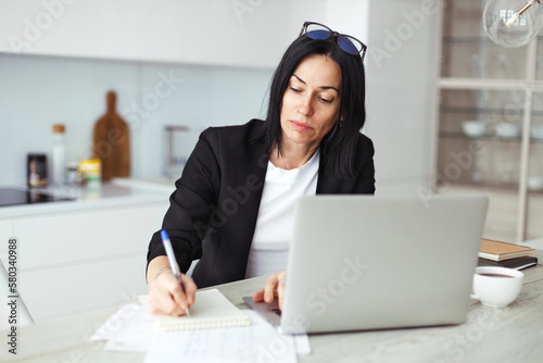 Female executive manager or accountant general working at kitchen table sitting in front of laptop, writing information or schedule in notebook, wearing glasses on head, dressed in jacket