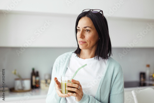 Good-looking pretty brunette female of 50 in casual home clothes with glasses on head standing at kitchen with transparent cup of celery juice in hands with green straw  going to enjoy delicious drink