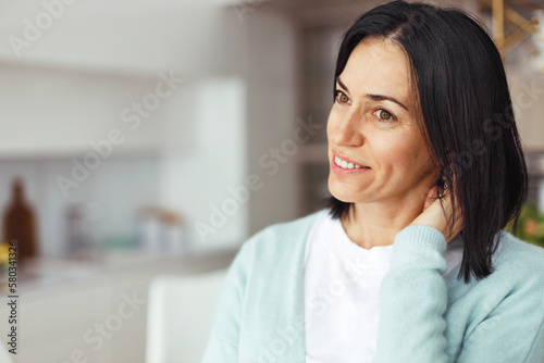 Portrait of shy charming mature brunette lady in blue cardigan standing in blurred home interior looking aside on copy space with smile, touching neck, feeling embarrassed and in love
