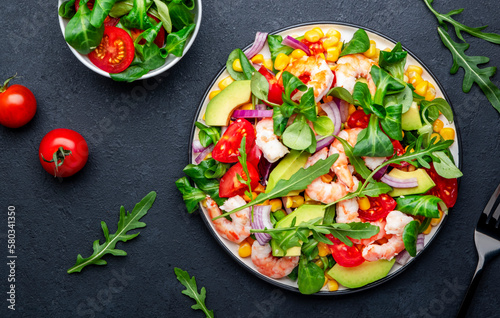 Delicious shrimp salad with sweet corn, avocado, cherry tomatoes, lamb lettuce and red onion on black table background. Top view