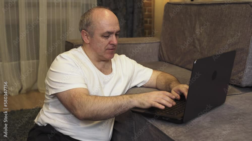 A mature man sits on the floor and works on a laptop that is on the sofa. A mature man sits on the floor and works on a laptop that is on the sofa.