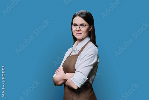 Portrait of confident young woman worker in apron on blue background