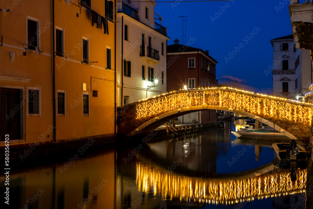 Chioggia cityscape in Venice Lagoon with narrow Vena water canal with colorful colorful boats among old buildings and illuminated brick bridge, blue evening sky, Veneto region, northern Italy