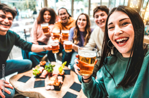 Girl taking a selfie while toasting beers - Friends clinking ale at brewery bar indoor at patio party - Friendship concept with young people having fun together drinking at happy hour promotion .